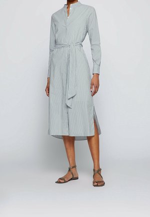 Femme portant une robe blanche à manches longues et longueur genoux, avec des rayures noires verticales, une ceinture à la taille, des fentes sur les côtés et des sandales à lanières vert olive.