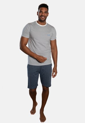 Smiling man with short dark hair wearing a striped white and black t-shirt and dark gray shorts, standing barefoot against a plain background.