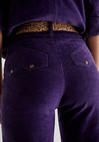 I pantaloni di velluto a coste viola presentano un design a righe testurizzato, arricchito da due tasche posteriori e una cintura decorata in tonalità di marrone e oro.