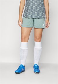 Shorts de sport verts, chaussettes blanches jusqu'aux genoux et crampons de football bleus. La tenue présente un haut à motif géométrique dans des tons de gris.
