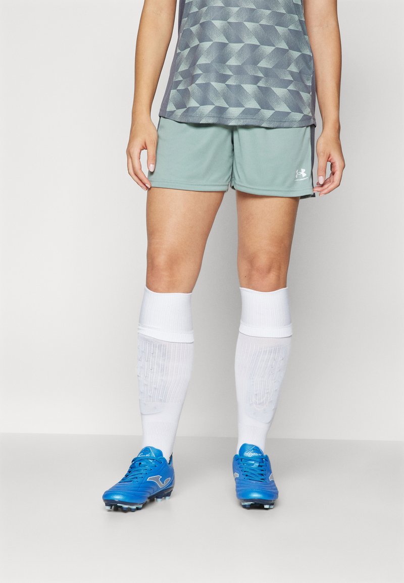 Shorts de sport verts, chaussettes blanches jusqu'aux genoux et crampons de football bleus. La tenue présente un haut à motif géométrique dans des tons de gris.