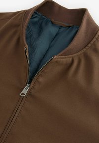 Braune Bomberjacke mit glatter Außenseite, geripptem Kragen und Reißverschluss. Das Innenfutter hat eine strukturierte blaue Fütterung mit geometrischen Mustern.