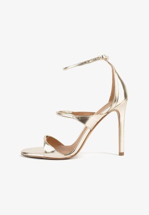 Kuld metallik kõrge kontsaga sandaal õhukese pahkluu rihmaga, avatud varvastega ja mitme rihmaga üle jala; sile tekstuur ja stiilne disain.