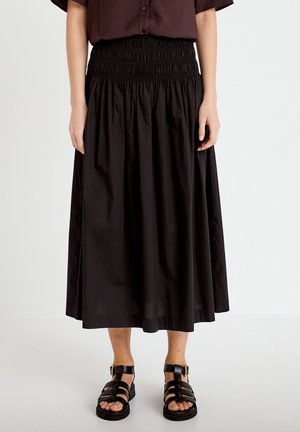 Donna che indossa una lunga gonna nera arricciata, camicia scura a maniche corte e sandali neri con cinturini, in piedi davanti a uno sfondo bianco.