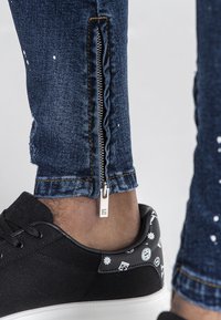 Jeans de mezclilla azul oscuro con detalle de cremallera lateral y desgastes, combinados con zapatillas de lona negras con acentos gráficos blancos.