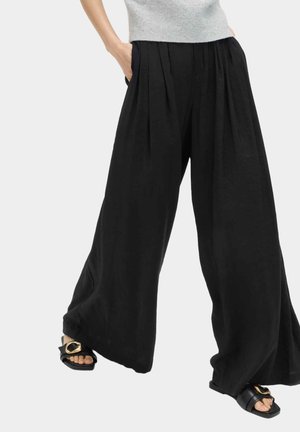 Osoba nosząca luźne, czarne spodnie o szerokich nogawkach z plisami, połączone z czarnymi sandałami z złotymi klamrami.