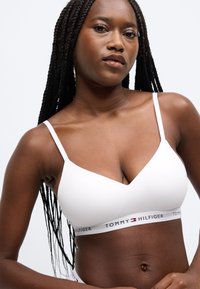 Bielá bezdrôtová podprsenka s výstrihom do V, tenkými ramienkami a elastickou páskou s logom v červenej, bielej a modrej farbe.