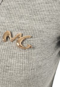Il tessuto a righe grigio presenta un accento di una spilla con logo "MC" dorato sul lato sinistro. La texture è morbida e leggermente strutturata.