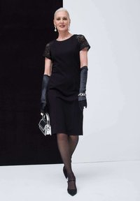 Schwarzes Kleid mit Spitzenärmeln, kombiniert mit langen schwarzen Handschuhen und einer kleinen silbernen Handtasche. Präsentation von spitzen schwarzen Absätzen und durchsichtigen Strumpfhosen.