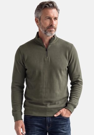 Mann mit grauen Haaren und Bart, der einen olivgrünen Langarmpullover mit Halbreißverschlusskragen und dunkelblaue Jeans trägt, steht mit einer Hand in der Tasche.