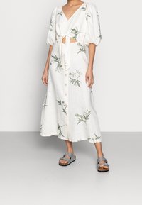 Robe en lin blanc avec manches courtes bouffantes, fermeture à nouer sur le devant, imprimé floral et fermeture à boutons ; associée à des sandales grises à enfiler.