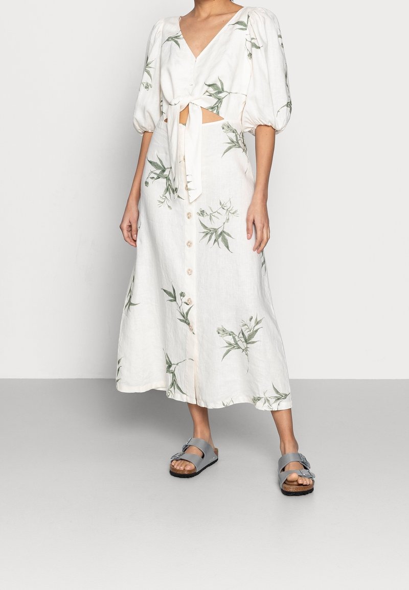 Robe en lin blanc avec manches courtes bouffantes, fermeture à nouer sur le devant, imprimé floral et fermeture à boutons ; associée à des sandales grises à enfiler.