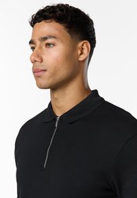 Jeune homme aux cheveux courts et bouclés portant une chemise noire texturée avec une fermeture éclair sur le devant, regardant vers la gauche sur un fond blanc.