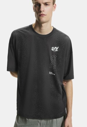 Fiatal férfi fekete, túlméretezett rövid ujjú ingben, rajta "RUN ON" és "Swiss Engineering" felirat, fehér háttér előtt áll.