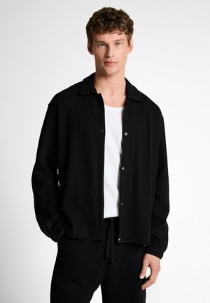 Junger Mann mit lockigem Haar, der eine schwarze, geknöpfte Jacke über einem weißen Hemd und passende schwarze Hosen vor einem schlichten Hintergrund trägt.