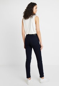 Dopasowane granatowe dżinsy z pięcioma kieszeniami, widocznym przeszyciem i lekkim streczem. W połączeniu z białą bluzką bez rękawów i białymi sneakersami.