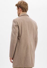Mann mit hellbraunen Haaren, der einen beigen Wollmantel und einen hellen Rollkragenpullover trägt, steht mit dem Rücken zu einem schlichten weißen Hintergrund.