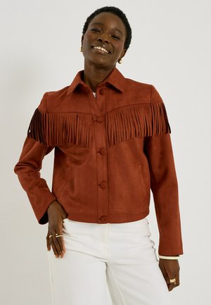 Mujer sonriente que lleva una chaqueta de ante color óxido con flecos en el pecho y los hombros y pantalones blancos, de pie con la mano en la cadera.