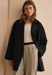 Svart oversized kappa med stora slag, texturerat tyg och en enkel knapp. Kombinerad med en ljus topp och glänsande, veckade byxor.
