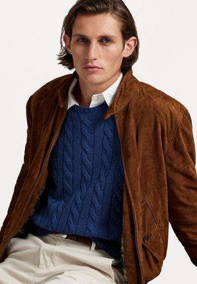 Καφέ σουέτ μπουφάν πάνω από ένα μπλε πουλόβερ με πλέξη καλωδίου, με λευκό πουκάμισο από κάτω. Το σύνολο διαθέτει υφασμάτινο σχέδιο και κουμπιά.