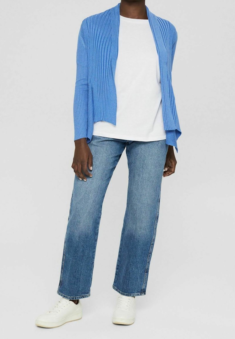 Personne portant un t-shirt blanc, un cardigan bleu côtelé ouvert, un jean droit bleu et des baskets blanches, debout devant un fond uni.