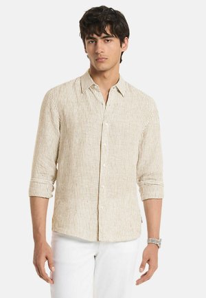 Ung mand iført en beige og hvid stribet langærmet skjorte med opkrogede ærmer og hvide bukser, stående mod en ensfarvet baggrund.