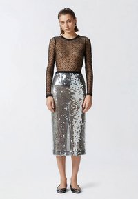 Juodos spalvos tinklinis ilgomis rankovėmis viršus su raštuotu dizainu, derinamas su sidabrine blizgančia pieštuko formos midi sijonu ir juodais lygiais batais iki kulkšnies.