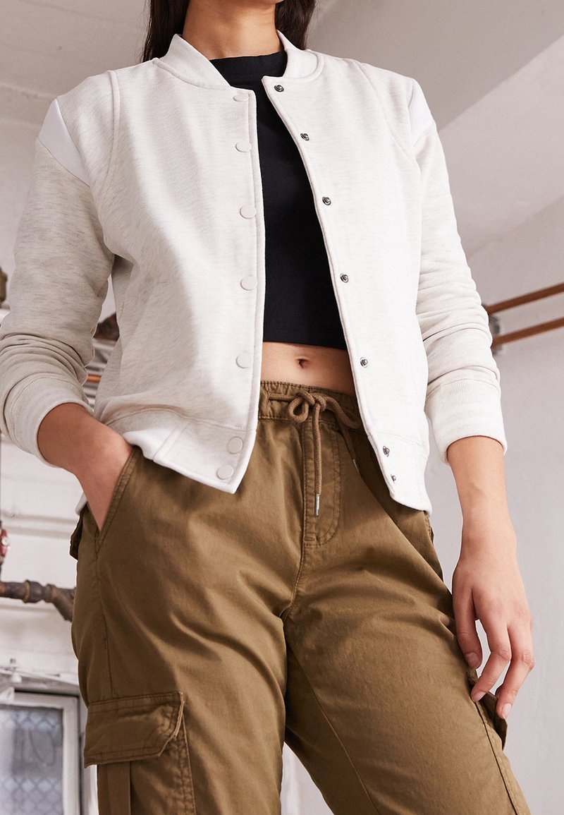 Femme portant une veste beige à boutons par-dessus un crop top noir et un pantalon cargo marron, debout les mains dans les poches dans un environnement intérieur.