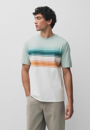 Nuori mies, jolla on kiharat tummat hiukset, pukeutuneena lyhythihaiseen raidalliseen t-paitaan ja vaaleansävyisiin housuihin, seisoo yksivärisen taustan edessä.