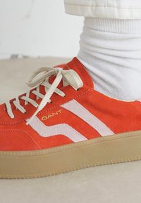 Piros bársony sneaker bézs fűzővel és talppal, fehér oldalsávokkal, fehér bordázott zoknival és világos színű nadrággal viselve.