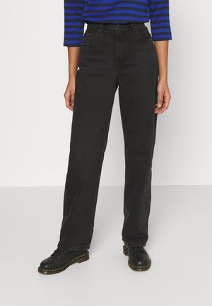 Persona con vaqueros negros rectos de talle alto, zapatos negros con cordones y una camiseta de manga larga a rayas azul y negra, de pie sobre un suelo gris.