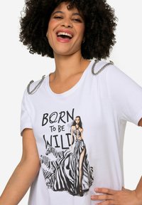 Hvid t-shirt med et sort-hvidt grafik af en model med zebra detaljer. Sølv kædedesign på skuldrene. Teksten siger "BORN TO BE WILD."