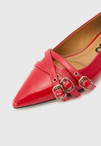 Chaussure rouge brillante à bout pointu avec deux sangles croisées dotées de boucles en argent, sur fond blanc uni.