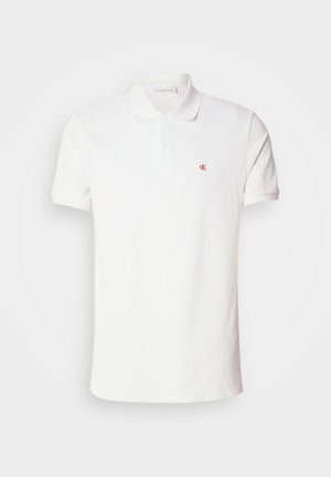 Polo blanc à manches courtes avec patte à deux boutons et petit logo rouge "CK" sur la poitrine gauche.