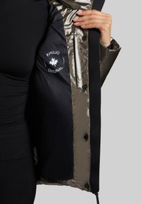 Polstret jakke med en mørk olivenfarvet overflade og sort inderside. Har et logo på indersiden af venstre side, trykknapper og lynlåslommer.