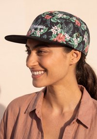 Mujer sonriente que lleva una gorra negra floral con hojas verdes y flores rosas, vestida con una camisa marrón claro con cuello.