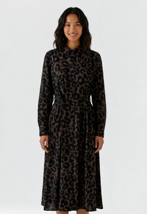 Vrouw draagt een midi-jurk met lange mouwen, knopen, luipaardprint, een kraag en een tailleceintuur, staand tegen een effen achtergrond.