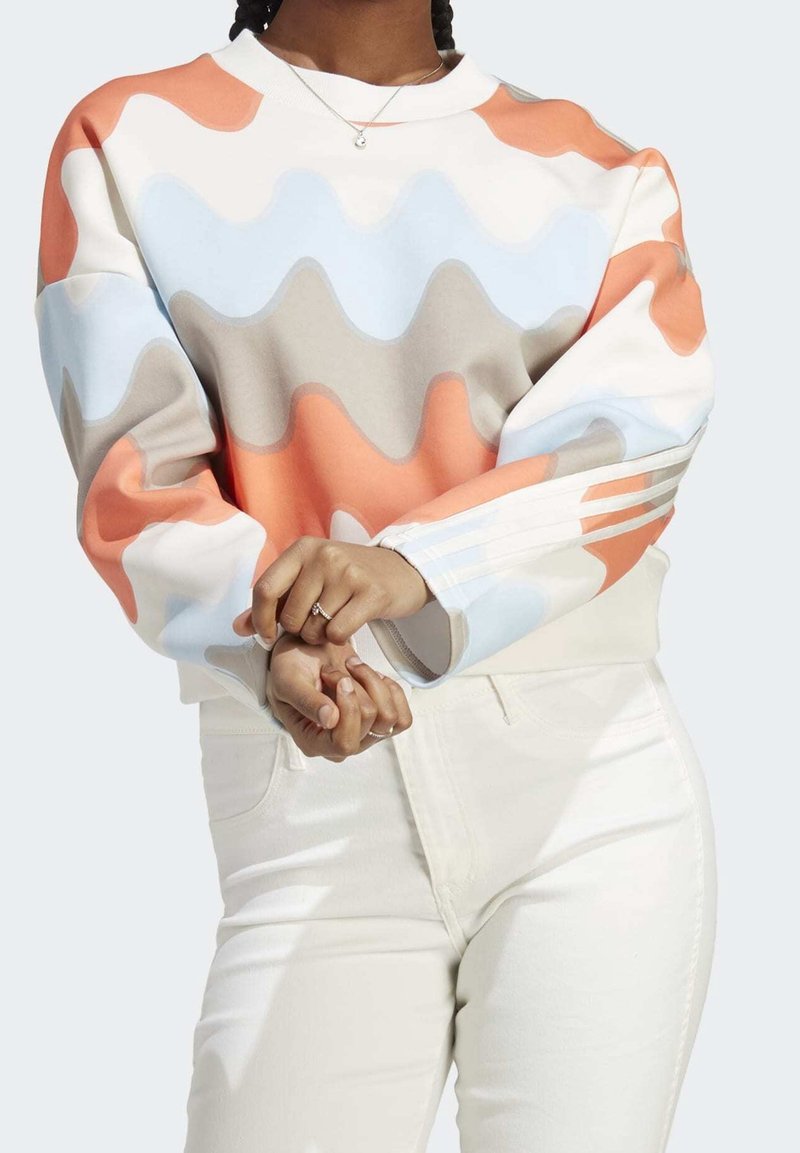 Sweatshirt à motif de vagues colorées dans des nuances d'orange, de bleu clair, de beige et de blanc. Équipé de poignets côtelés et d'une coupe décontractée. Associé à un jean blanc.