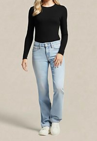 Femme portant un haut noir à manches longues, un jean bleu clair avec poches avant et des baskets blanches, debout devant un fond beige uni.