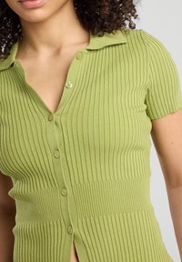 Short-sleeve, button-up top made of ribbed green knit fabric. Features a collar and slight side slits at the hem.