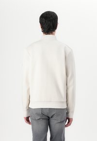 Helles beiges Sweatshirt mit hohem Kragen, gerippten Bündchen und Saum, glatter Textur und minimalistischer Gestaltung. Getragen über grauen Jeans.