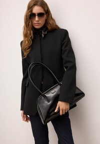 Sort læderhåndtaske med en glat tekstur, forlænget form og to runde håndtag. Har et sølvlogo-detalje på fronten.