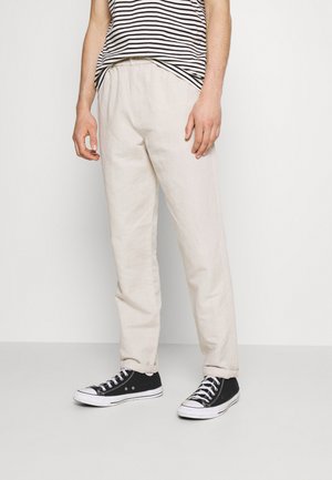 Uomo che indossa pantaloni beige, scarpe da ginnastica alte nere e una camicia a righe bianca e nera, in piedi contro uno sfondo bianco semplice.