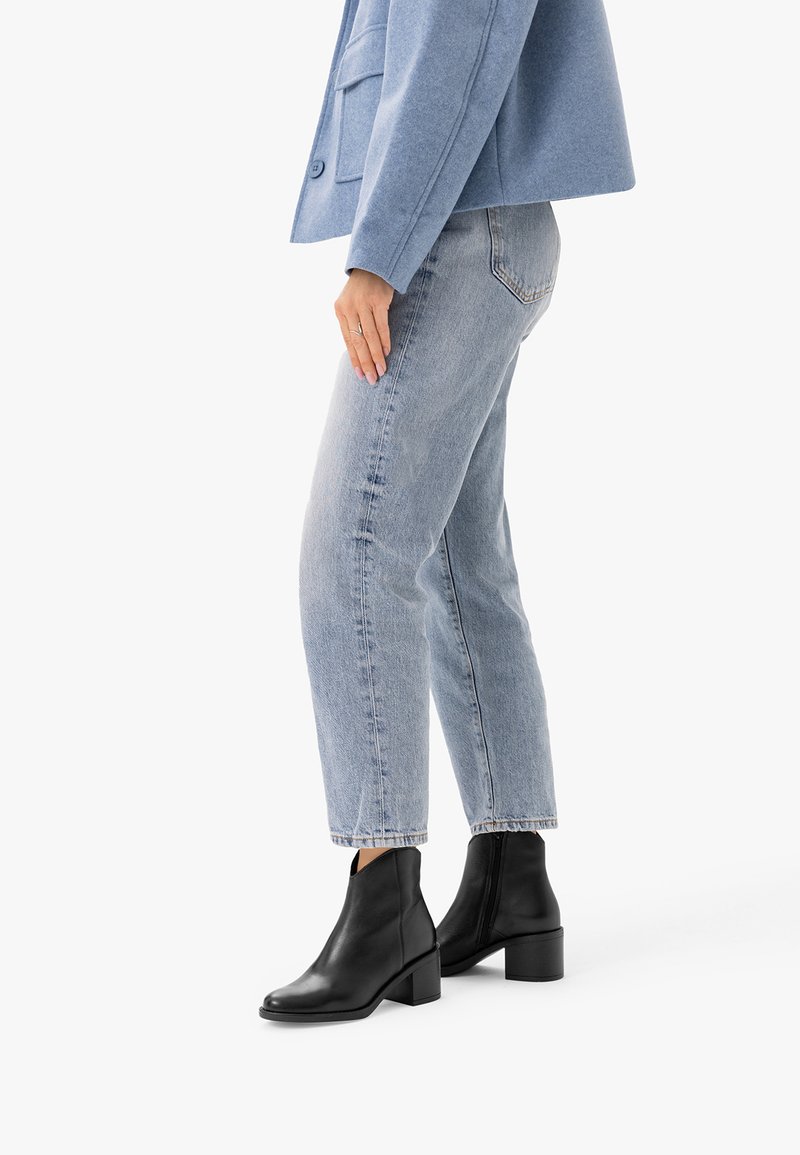 Personne portant un jean bleu clair, une veste bleue et des bottines noires, debout de profil sur un fond blanc.