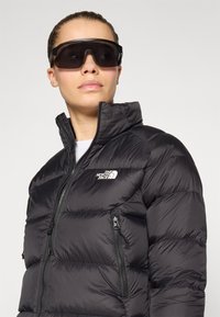 Čierna nafúknutá bunda s vysokým golierom, s većami na zips a logom The North Face. Tmavé veľké slnečné okuliare dopĺňajú vzhľad.