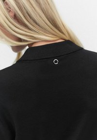 Mustassa neulepaidassa on ribbineulekaulus ja pieni hopeinen pyöreä logo selässä. Pehmeä rakenne ja istuva malli.