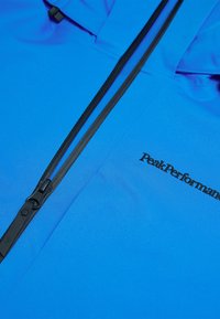 Blauwe jas gemaakt van lichtgewicht stof met een gladde textuur, voorzien van zwarte ritssluitingen en het "Peak Performance" logo in zwarte borduursels.