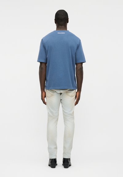 Άνδρας στέκεται με την πλάτη γυρισμένη, φοράει μπλε κοντομάνικο πουκάμισο και τζιν ανοιχτού χρώματος με μαύρα παπούτσια, σε απλό φόντο.