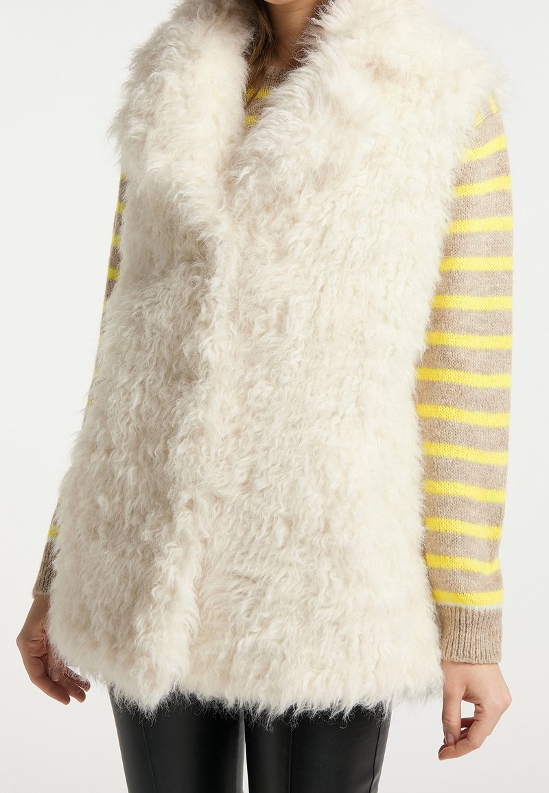 Biały kamizelka z sztucznego futra z głębokim kołnierzem i otwartym przodem, noszona na paskowanej bluzce, charakteryzująca się miękką teksturą i warstwowym wyglądem.