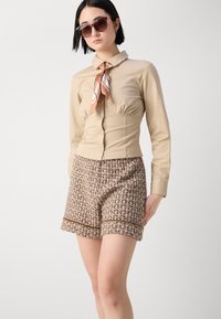 Beige, fitted shirt with a collar and tied scarf. Brown, textured tweed shorts with a checkered pattern and a hem accent. Black sunglasses.